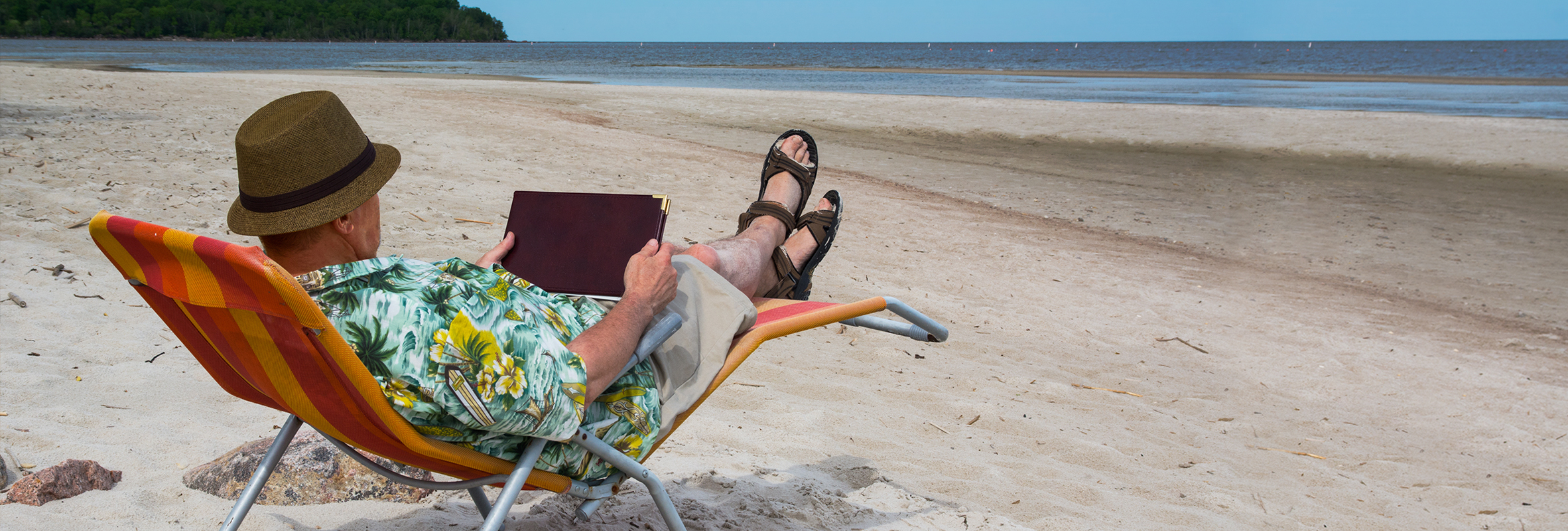 A retired man sitting on a chair on a sandy beach, holding a book and enjoying the sun.